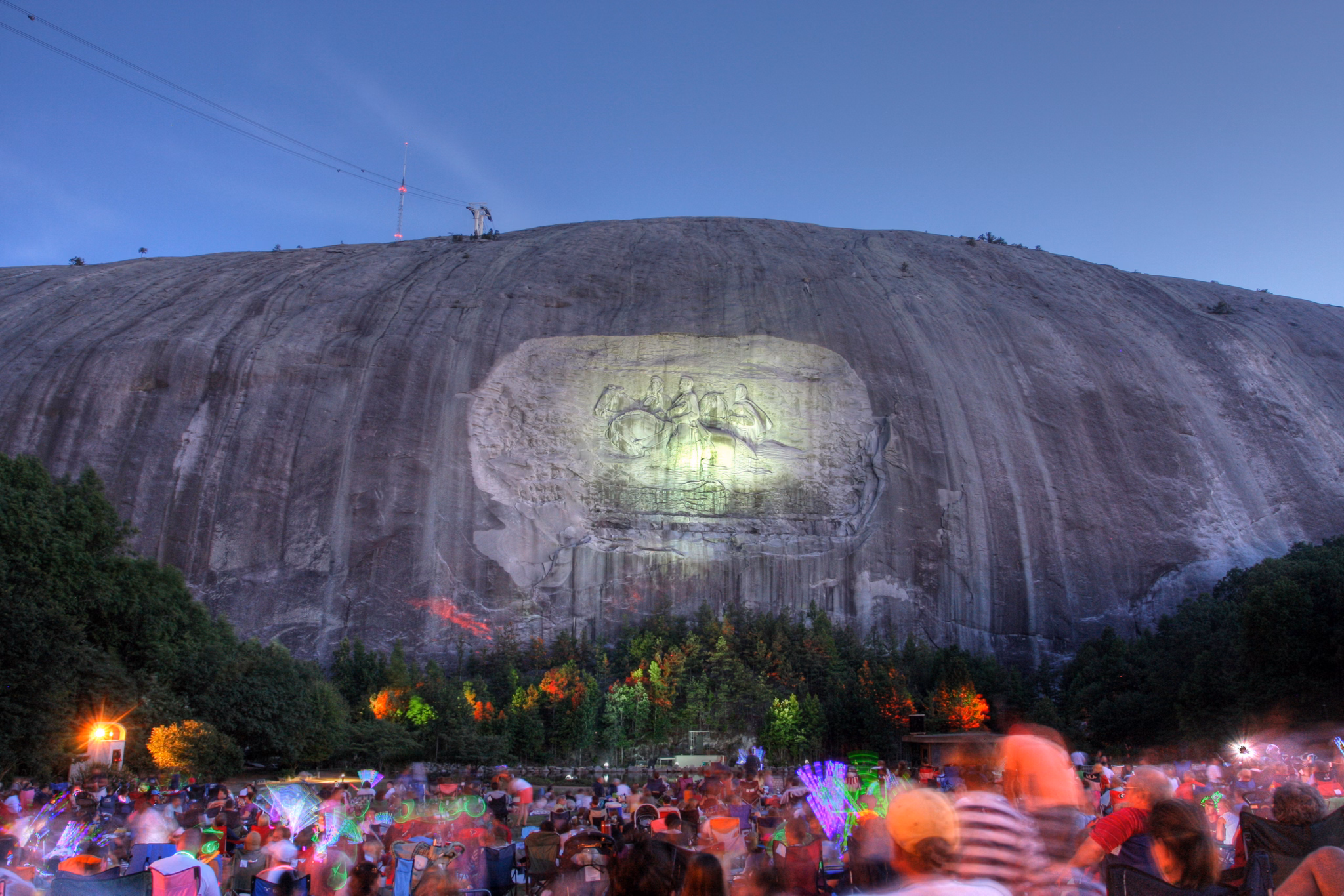 Illuminated carving on Stone Mountain, GA with nighttime crowd below holding colorful glowsticks during a special event.