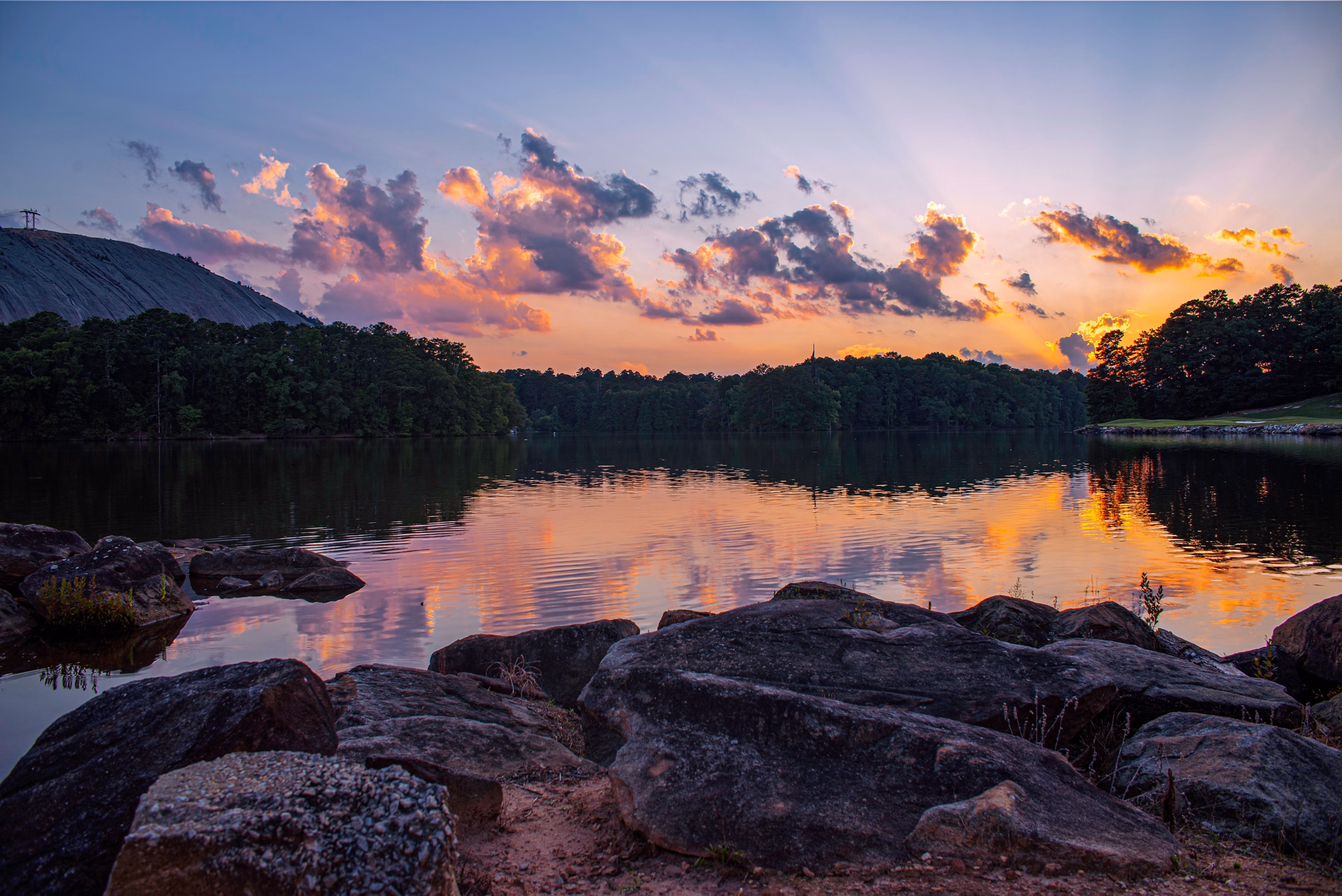 Sunset over lake in Dekalb County, GA with pink-orange clouds reflecting on calm water, rocky shoreline, and Stone Mountain.