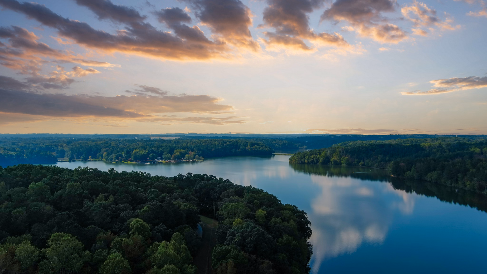 Aerial view of serene lake in Lithia Springs, GA at sunset, surrounded by dense forest with clouds reflecting on calm water.