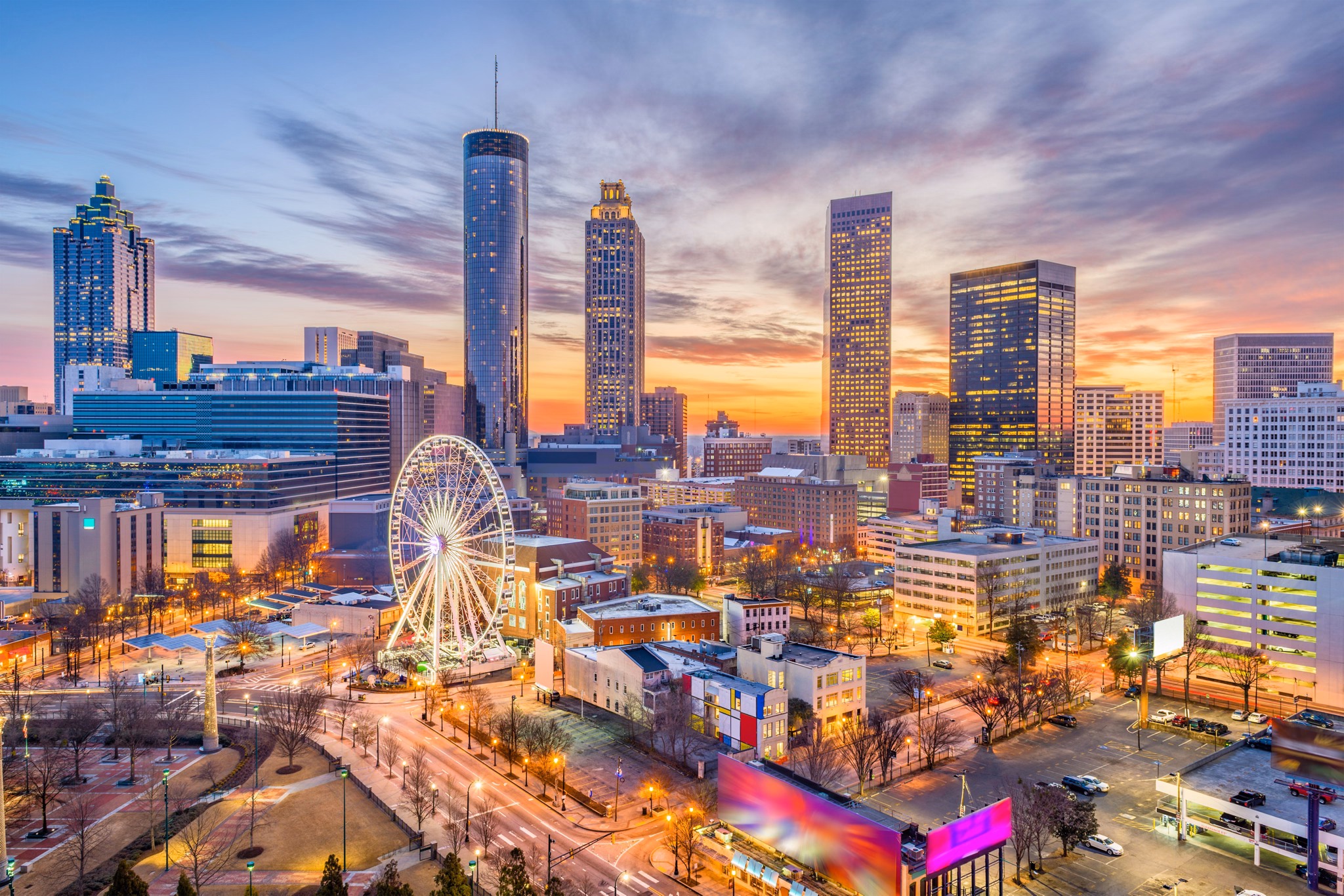 Downtown Atlanta, GA skyline at sunset with lit skyscrapers, Ferris wheel, and Centennial Park under colorful evening sky.