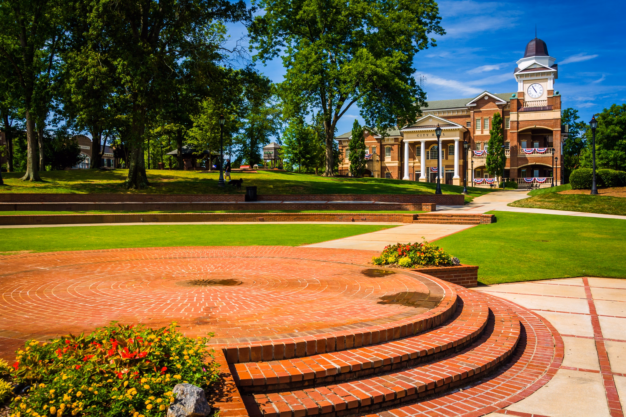 Brick plaza leading to Duluth, GA city hall with clock tower, American flag decor, and landscaped grounds with shade trees.