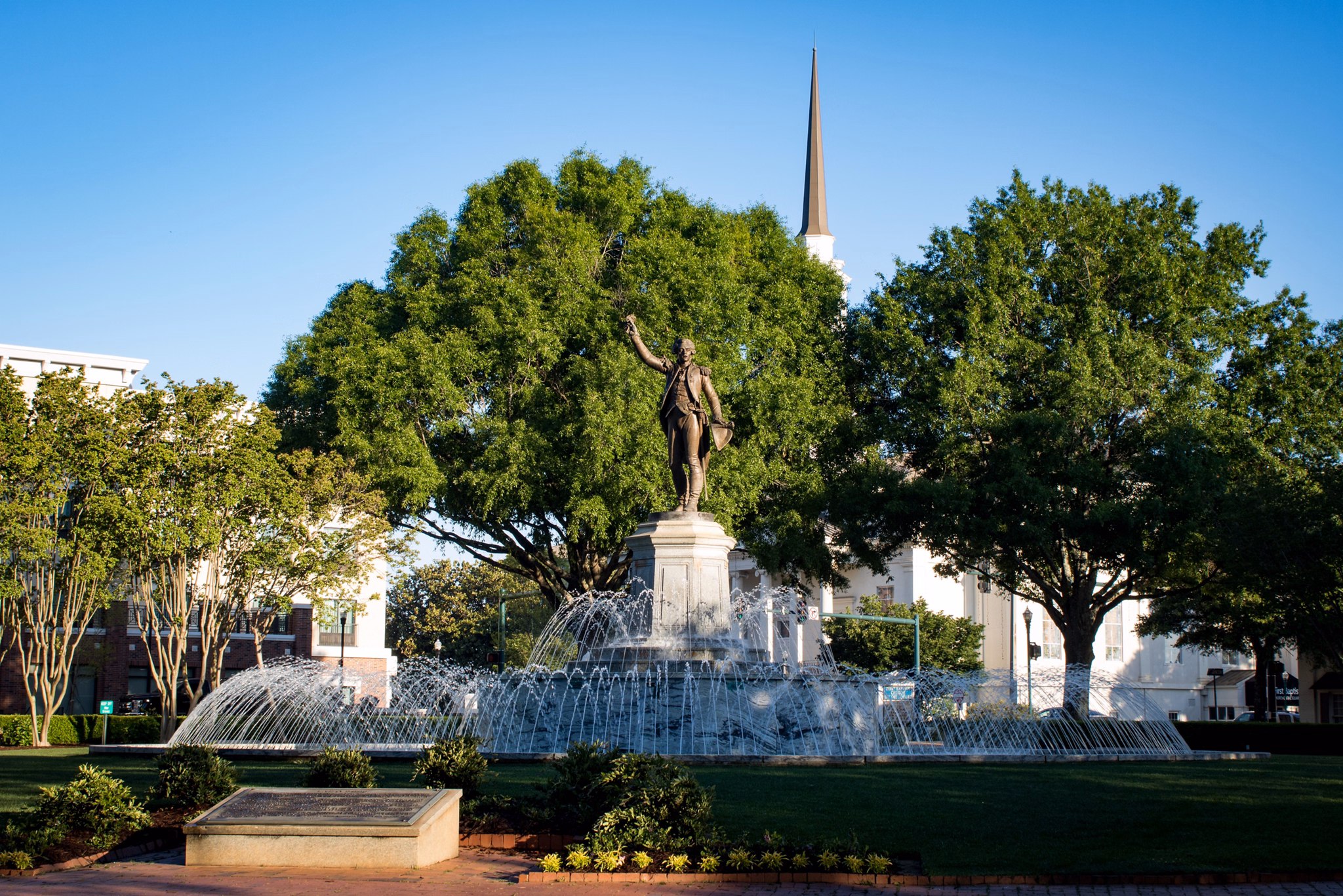 Fountain plaza in Lagrange/Newnan, GA with a statue of a historical figure, water jets, and a church steeple beyond trees.