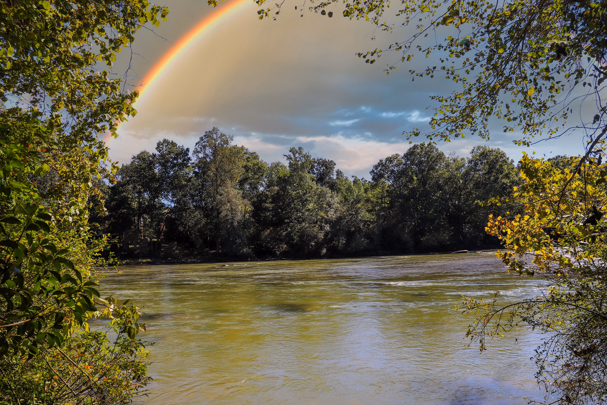 Rainbow arching over the Chattahoochee River in Johns Creek, GA with leafy trees framing the view of flowing water.
