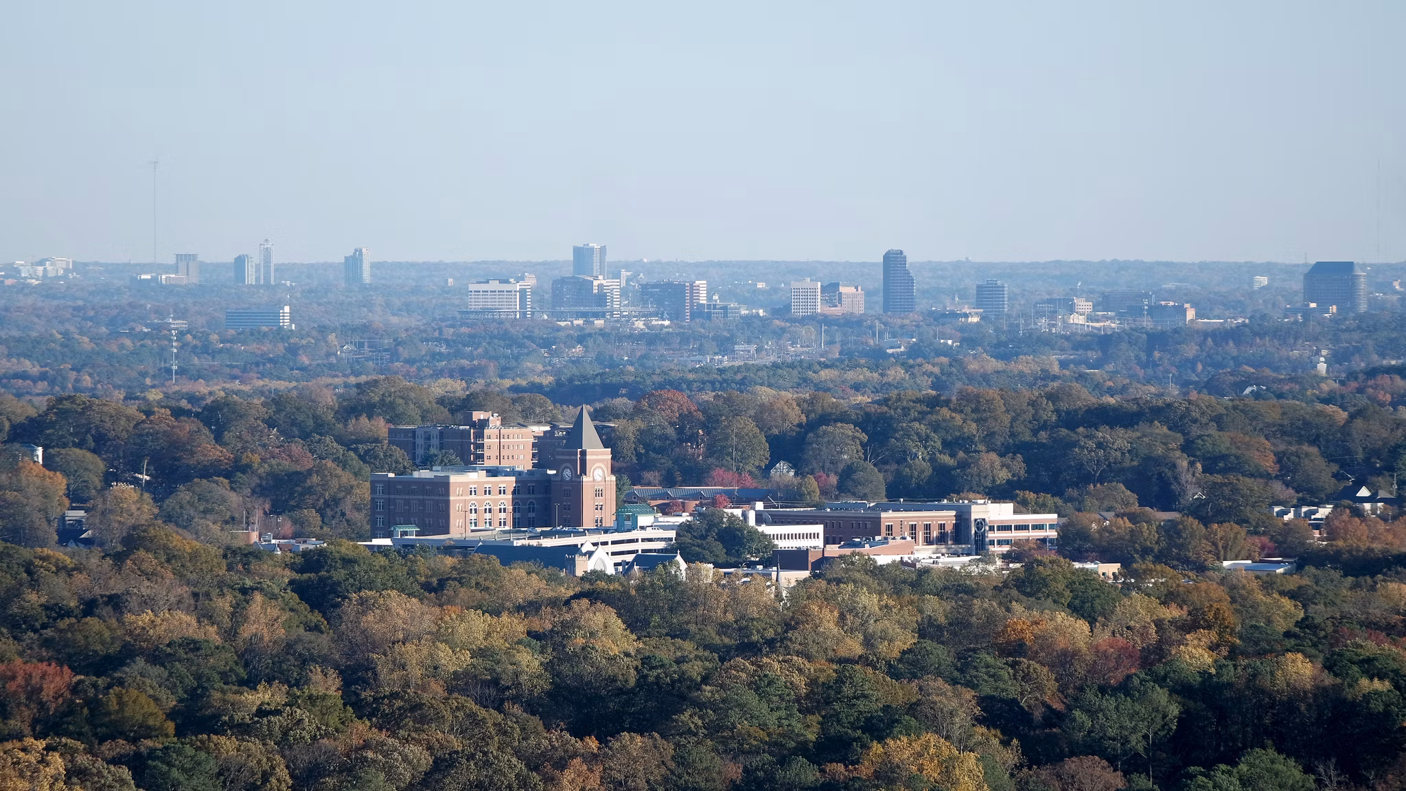Aerial view of Sandtown, GA with brick buildings, a clock tower, and autumn trees with the Atlanta skyline in the distance.