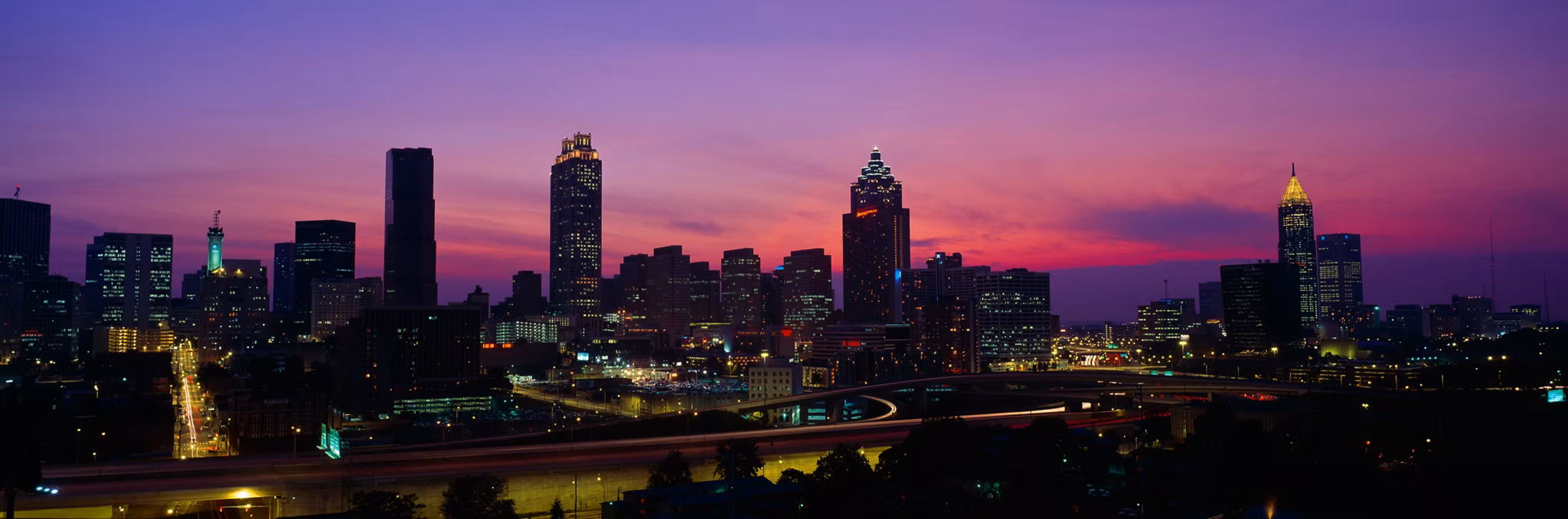 City skyline of Atlanta at sunset viewed from South Fulton County, GA, with purple and pink sky and illuminated skyscrapers.