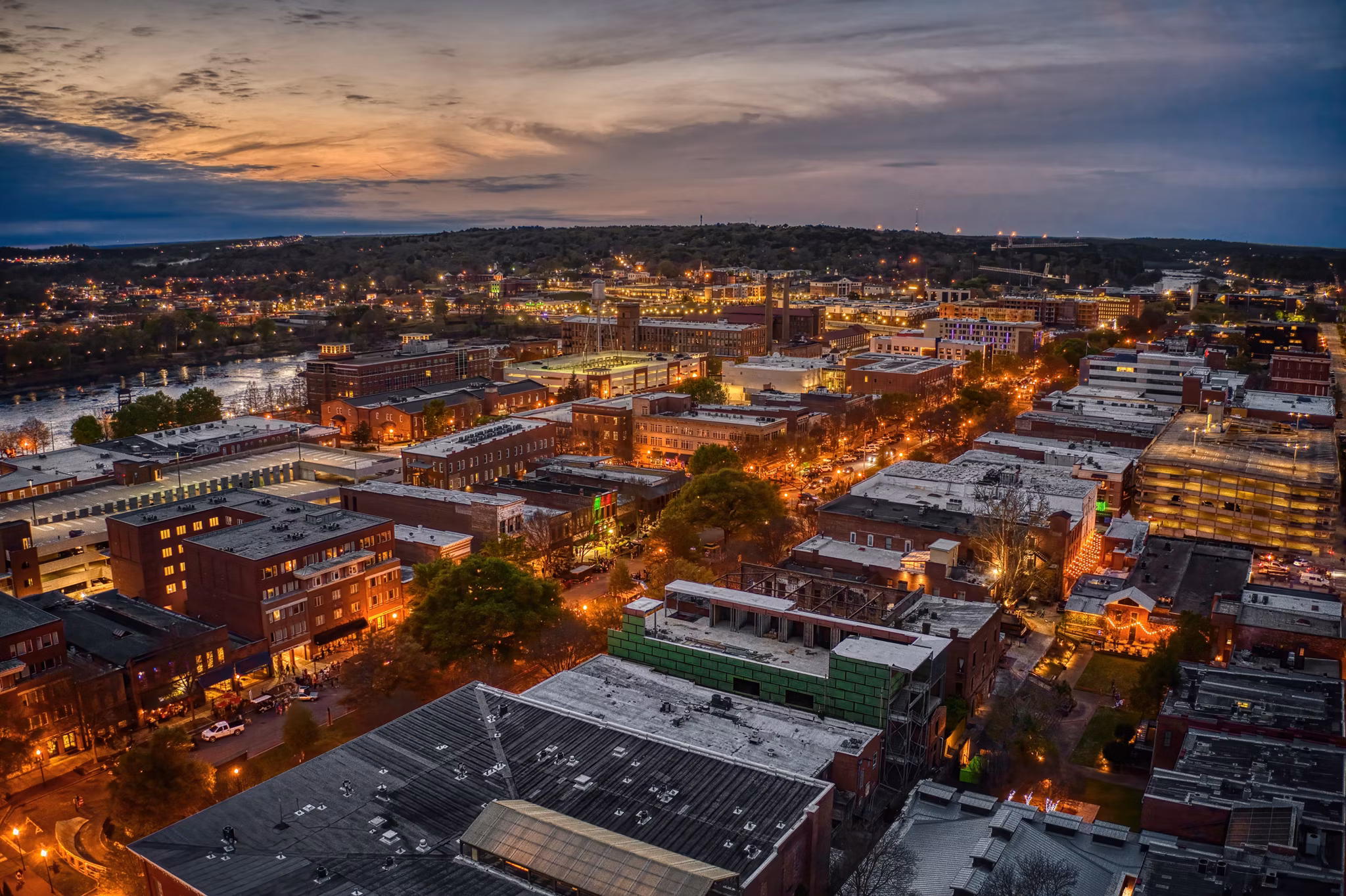 Night view of downtown Columbus, GA with illuminated brick buildings, city streets, and Chattahoochee River visible at dusk.