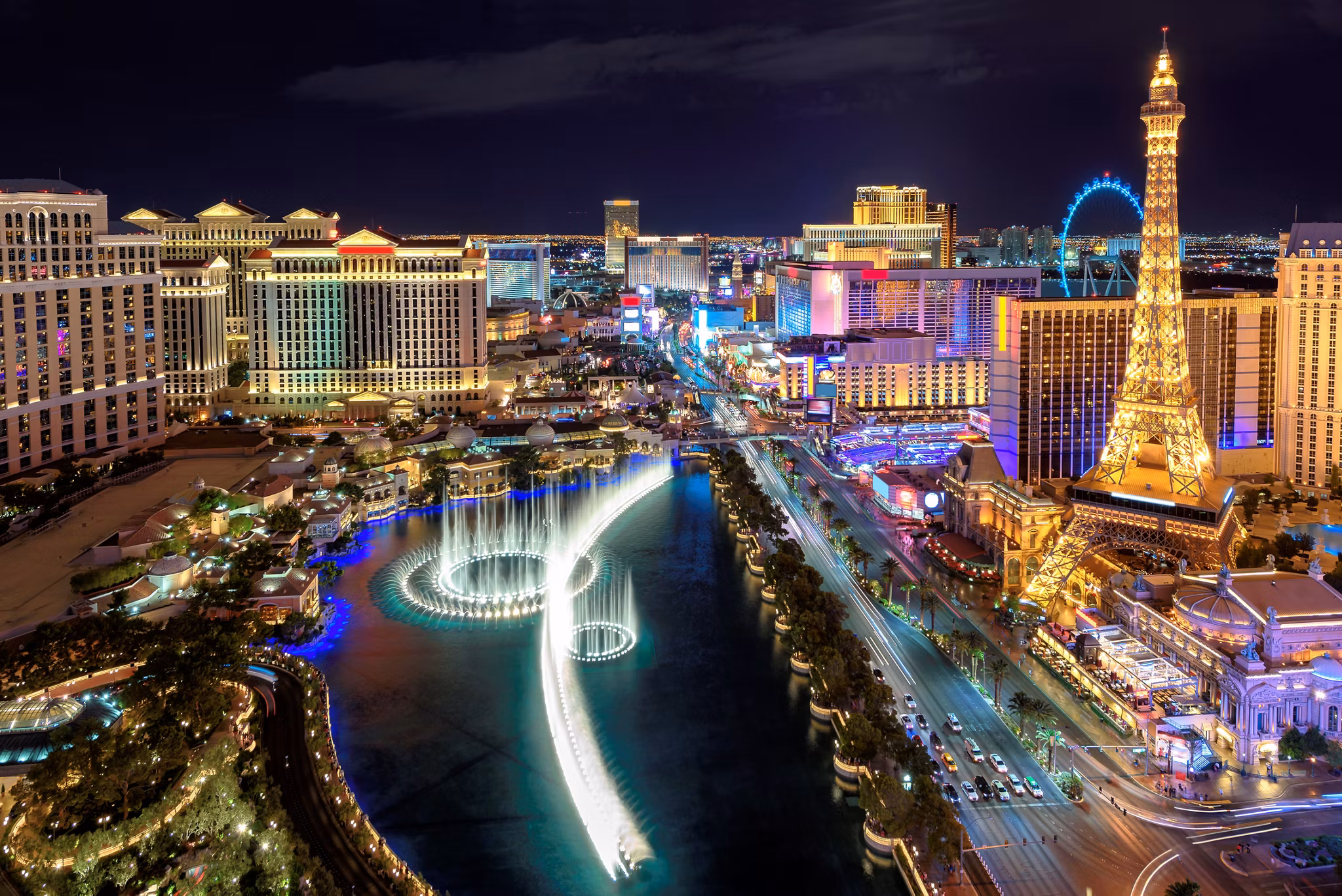 Aerial nighttime photograph of the Las Vegas Strip featuring the iconic illuminated Bellagio Fountains, Paris Las Vegas Eiffel Tower replica, High Roller observation wheel, and brightly lit casino resort hotels along the Strip