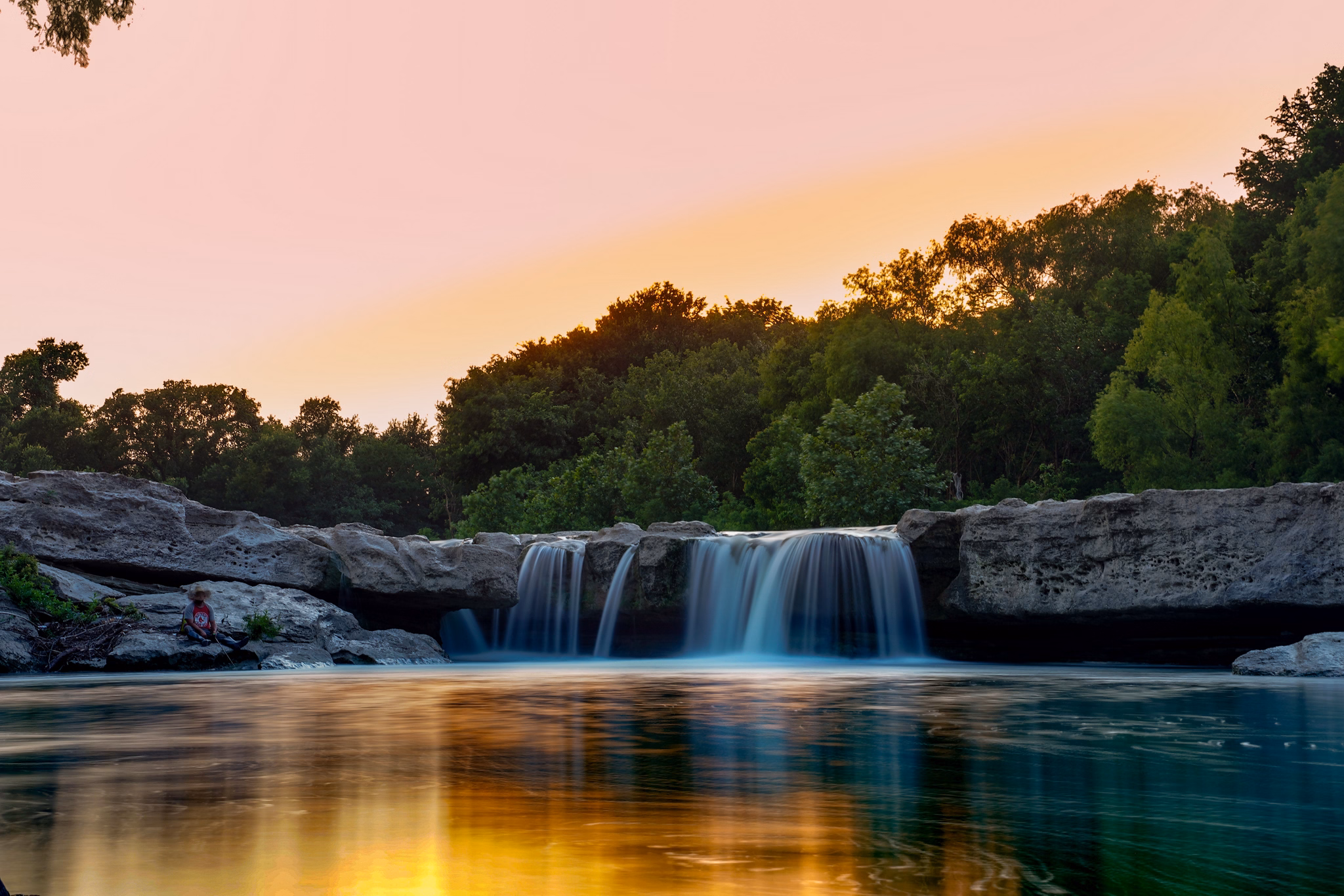 A peaceful waterfall in Bluff Springs/Onion Creek, Texas, surrounded by lush greenery under a colorful sky at sunset.
