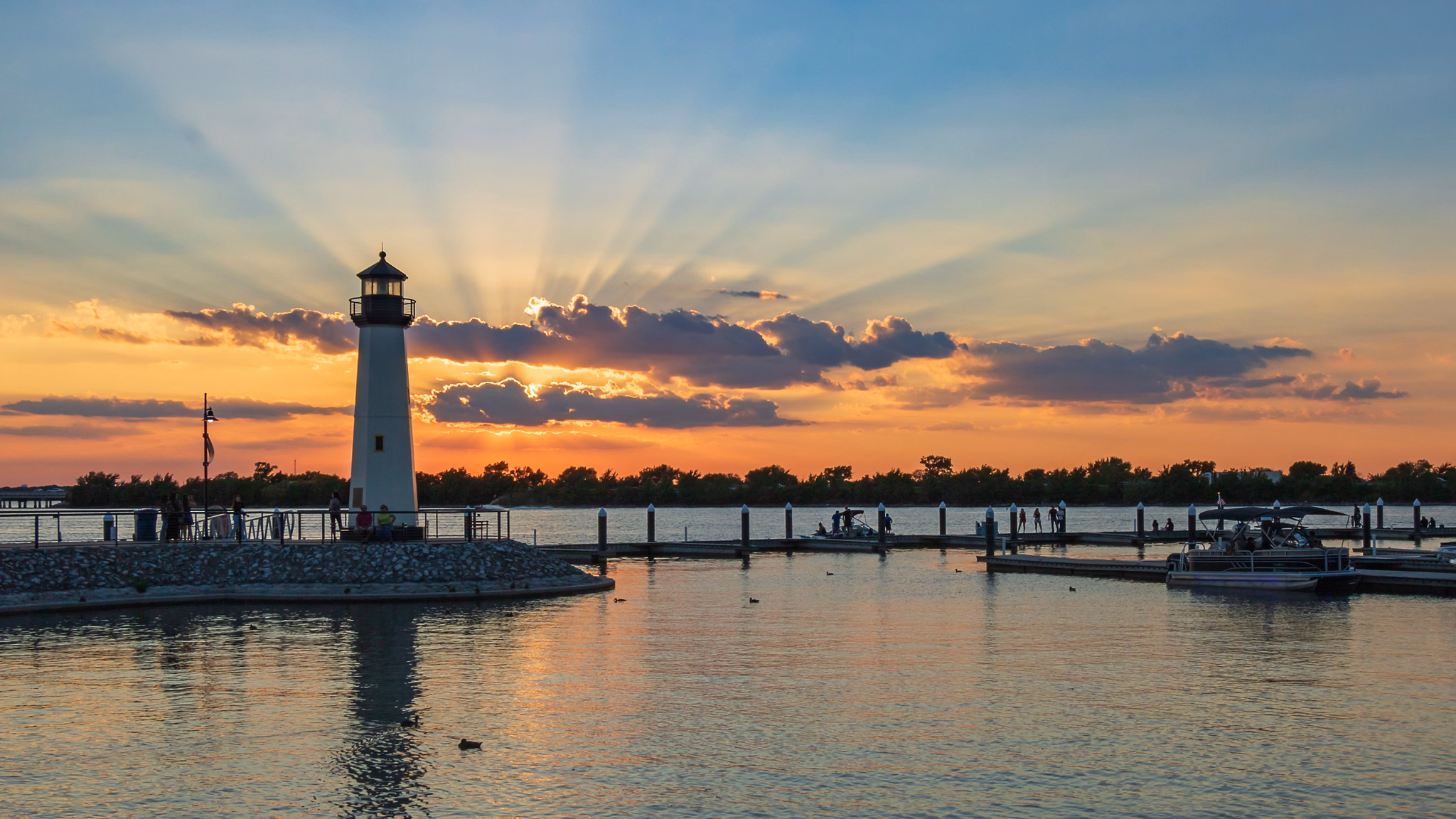 Sunset over a lake in Rockwall, Texas, with a lighthouse and people near the shore. The sky is filled with colorful clouds.