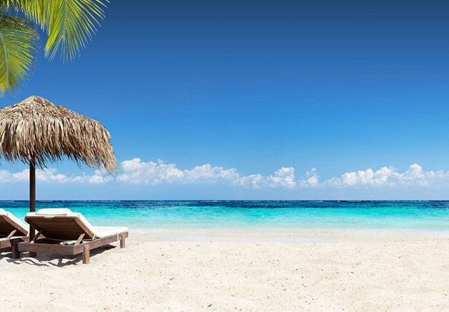 Tropical beach scene with clear blue sky, turquoise sea, white sand, a straw umbrella, and a pair of sun loungers.