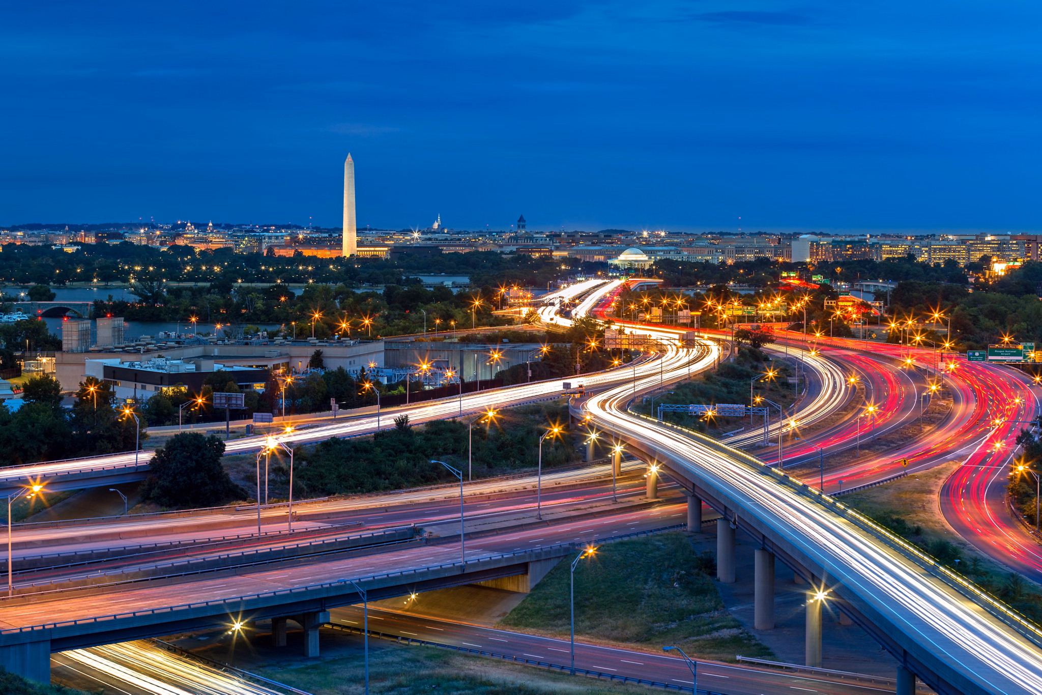 Washington D.C. cityscape at dusk with rush hour traffic trails on I-395 highway. Washington Monument, illuminated, dominates the skyline.