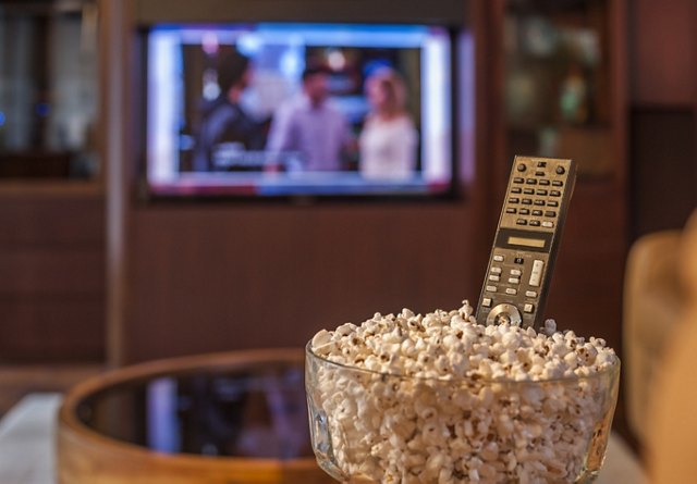 A bowl of popcorn and a remote control in the foreground with a blurred television screen in the background, suggesting a cozy movie night at home.