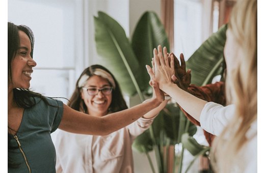 Three women smiling and giving a high-five, celebrating teamwork in a room with natural lighting and green plants.