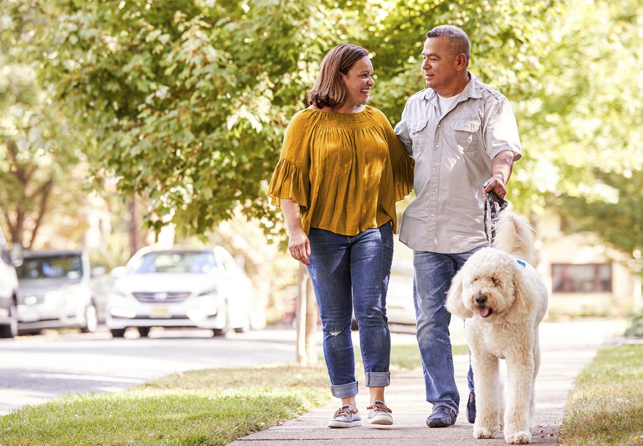 A smiling couple walking their fluffy white dog on a sunny suburban sidewalk.