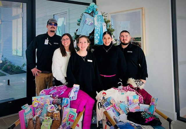 Group of five people smiling in front of a Christmas tree with various children's gifts.