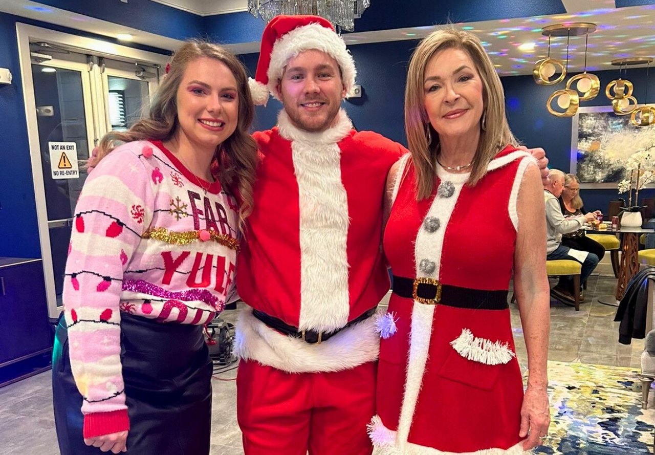 Three people in festive attire, including a man in a Santa Claus costume, smiling indoors.