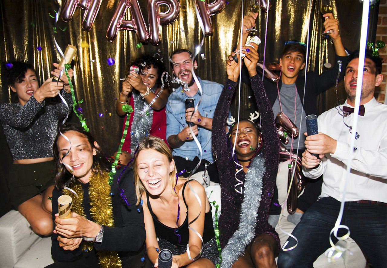 Energetic partygoers celebrating with party poppers, laughter, and 'Happy' balloons in the background.