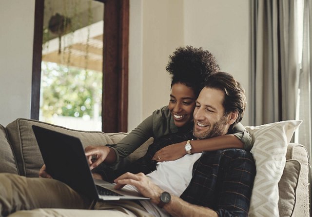 A couple relaxing on a couch with a laptop, smiling and enjoying each other's company.