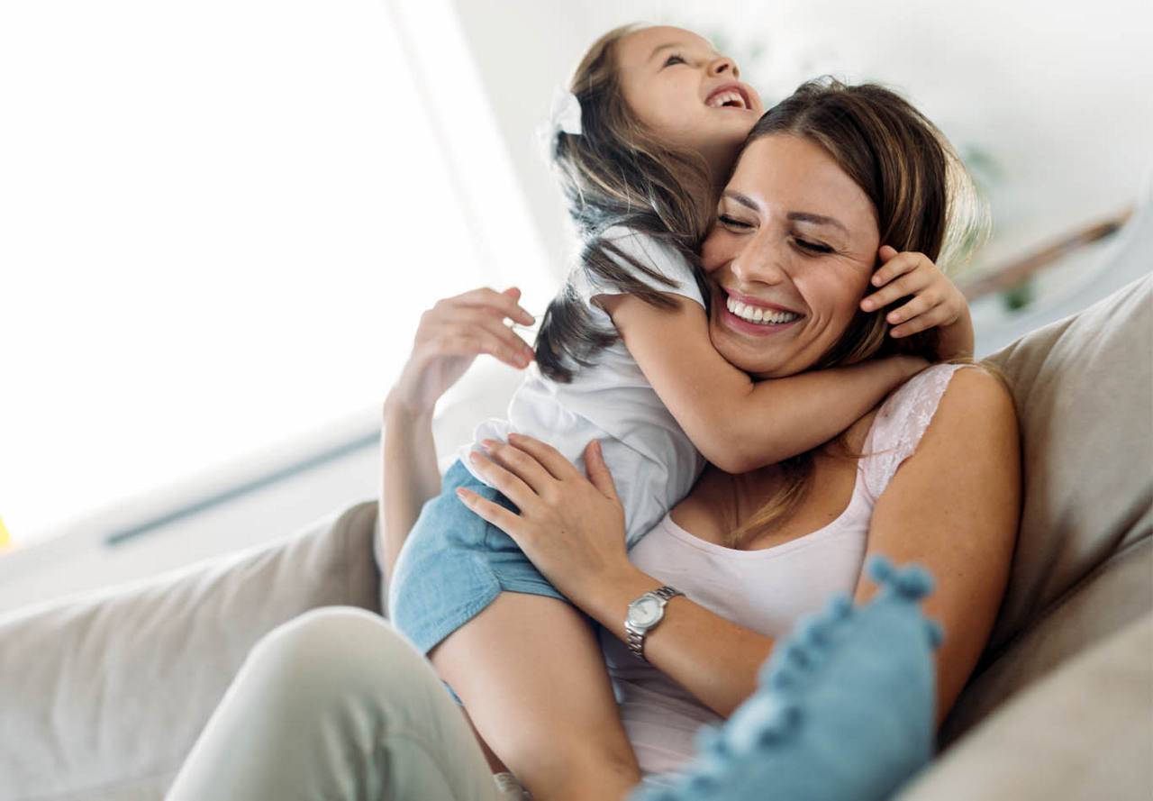 A young girl embracing her laughing mother from behind on a couch, displaying affection and happiness.