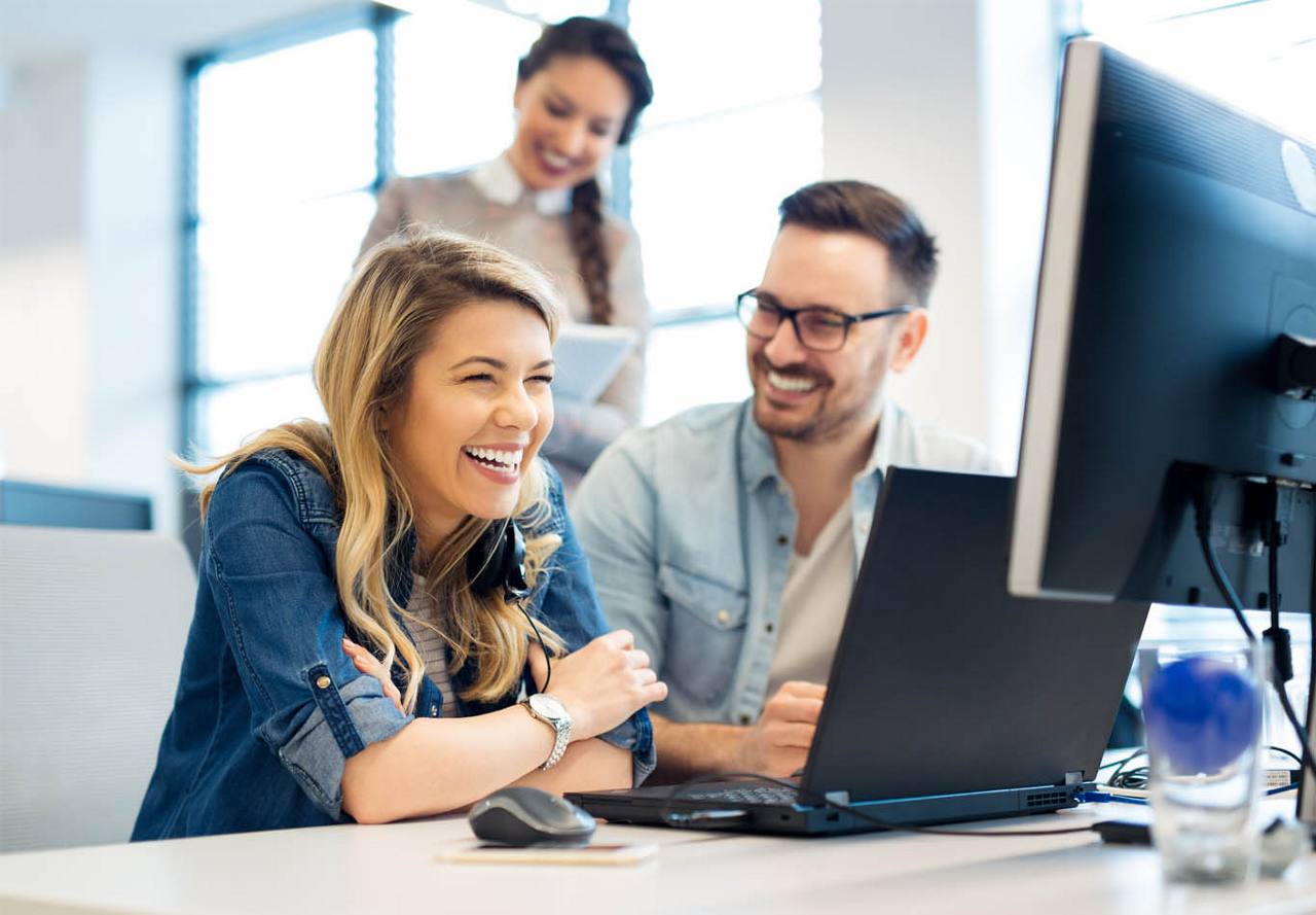 Two colleagues laughing together in a bright office setting, with another coworker in the background.