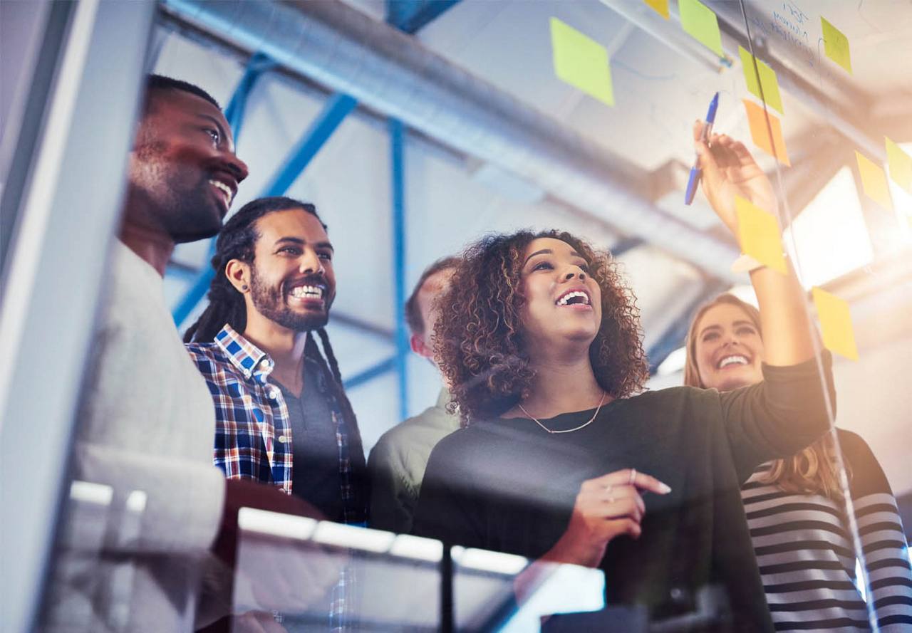 A team of smiling colleagues brainstorming with sticky notes on a glass wall in a bright office.