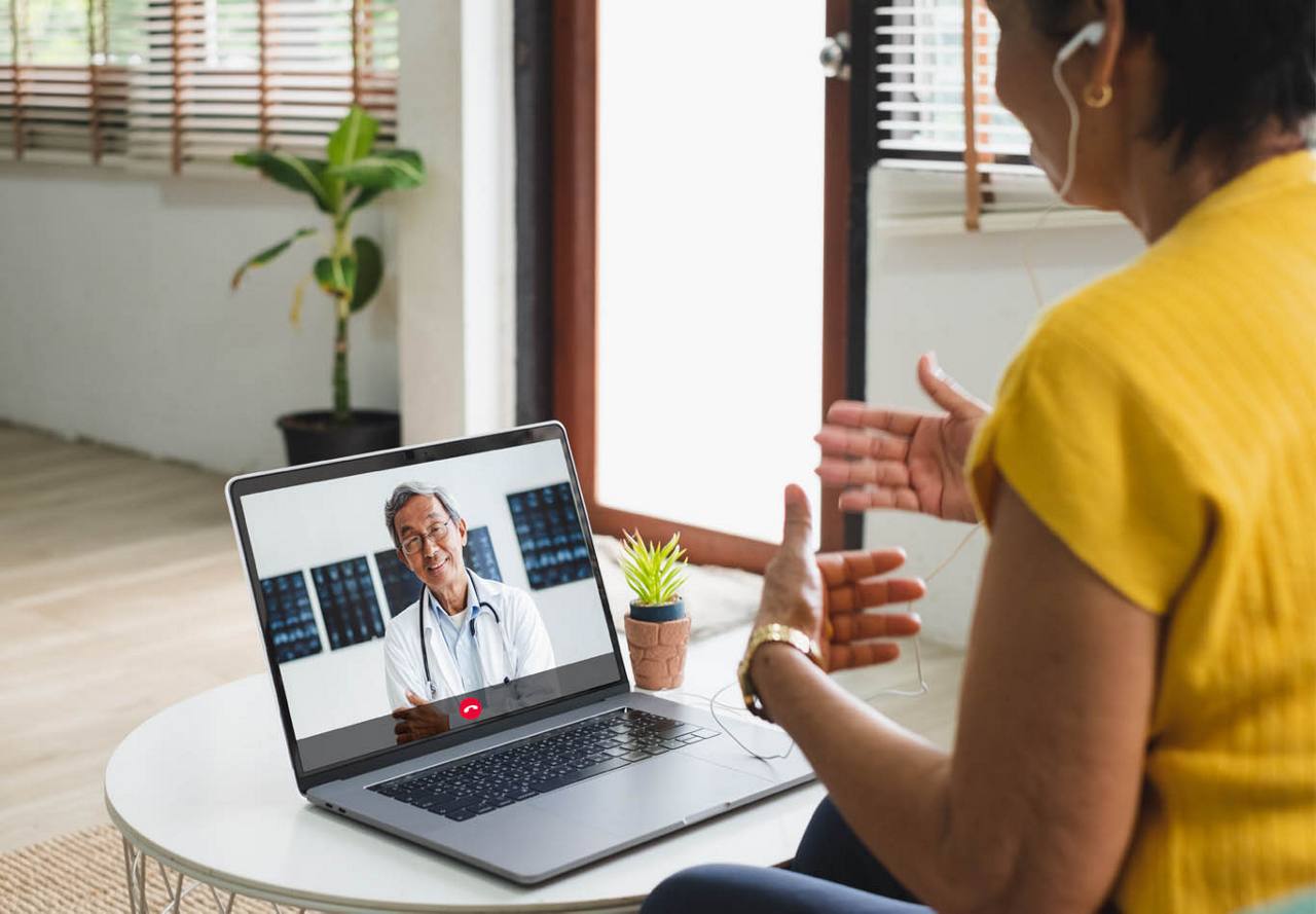 A woman in a yellow shirt having a video call with a smiling male doctor displayed on her laptop screen.