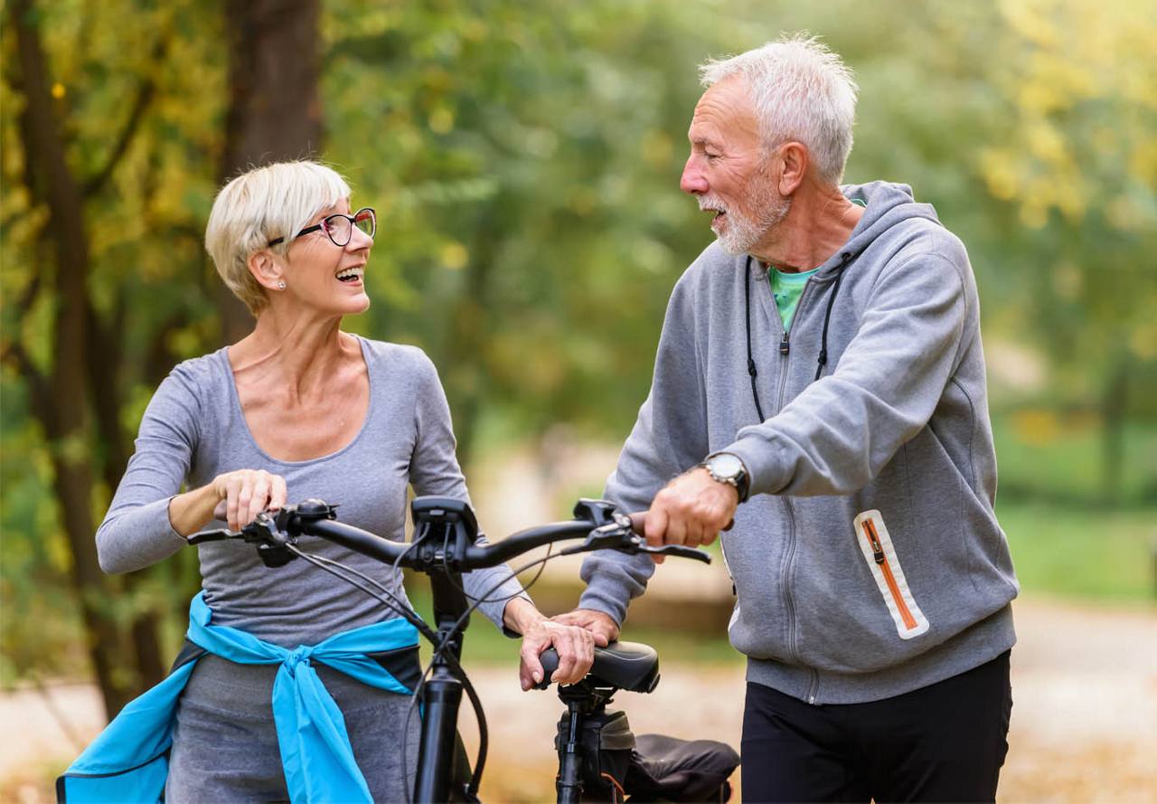 Senior couple joyfully talking and holding bicycles in a park, surrounded by trees.