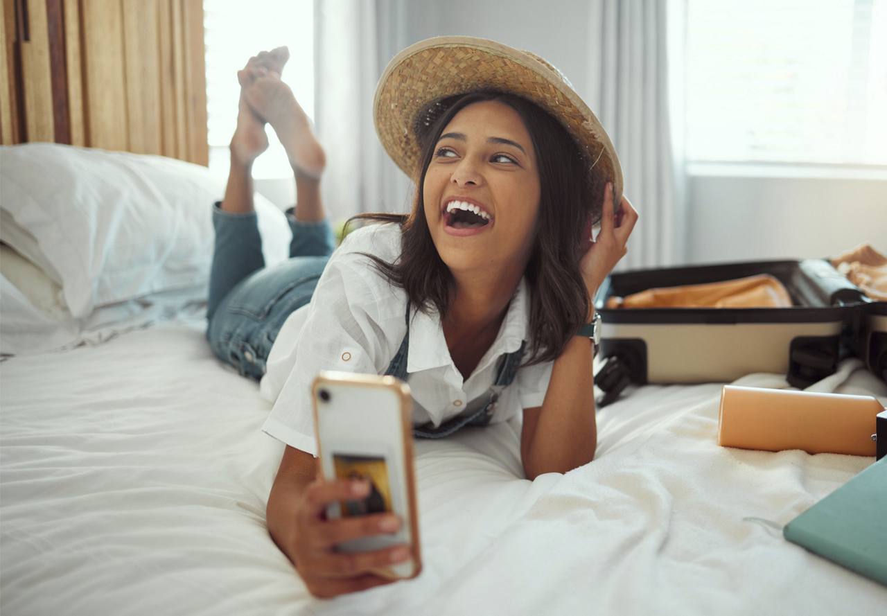 A joyful woman in a straw hat lying on a bed taking a selfie, with an open suitcase beside her.