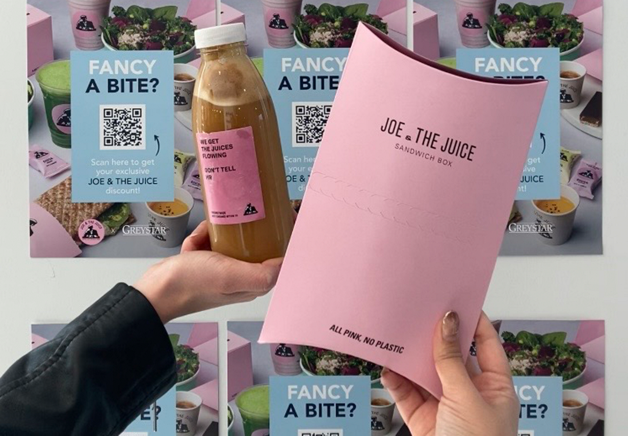 Hands holding a pink sandwich box and a bottle of juice against a promotional backdrop offering a discount.