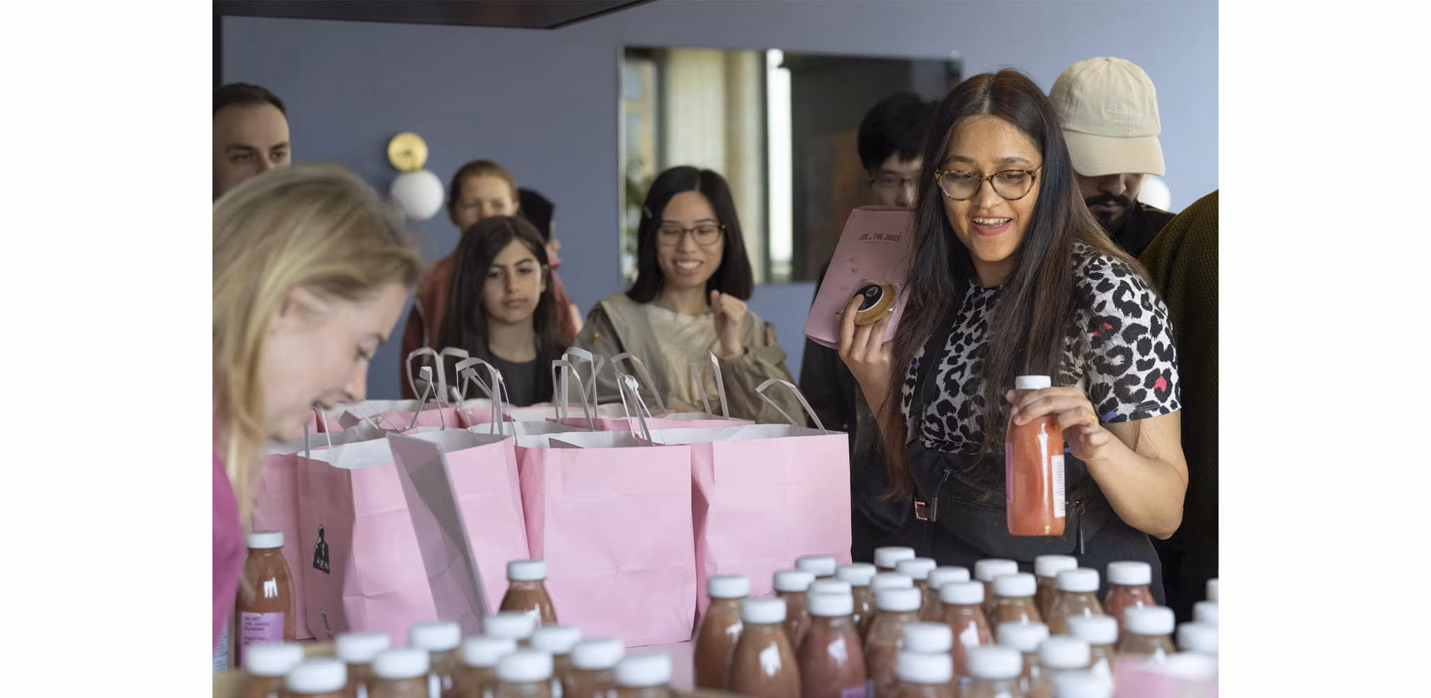 A diverse group of people happily gathering around a table with pink gift bags and bottles of juice.