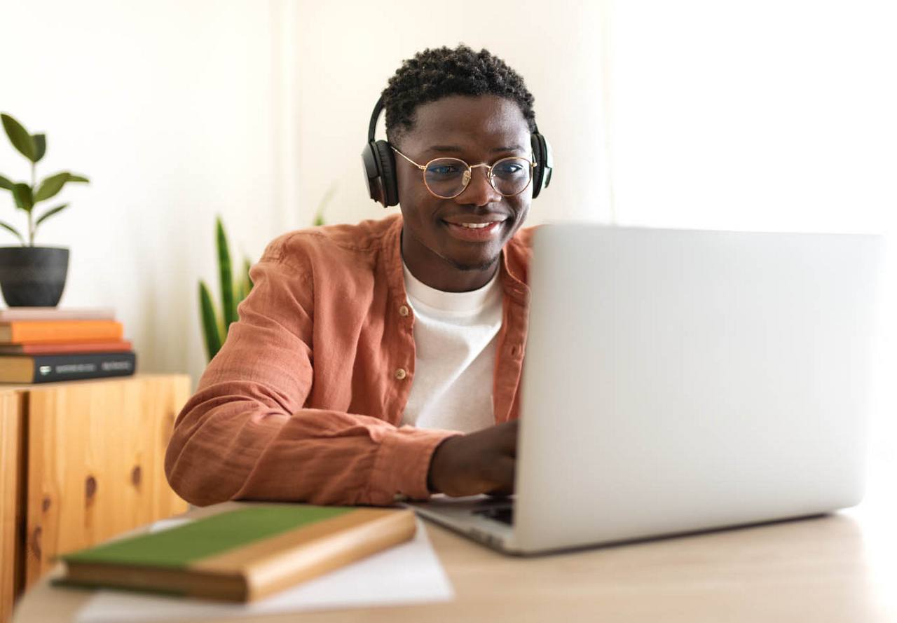Cheerful young man with headphones using a laptop in a bright room with plants and books in the background.