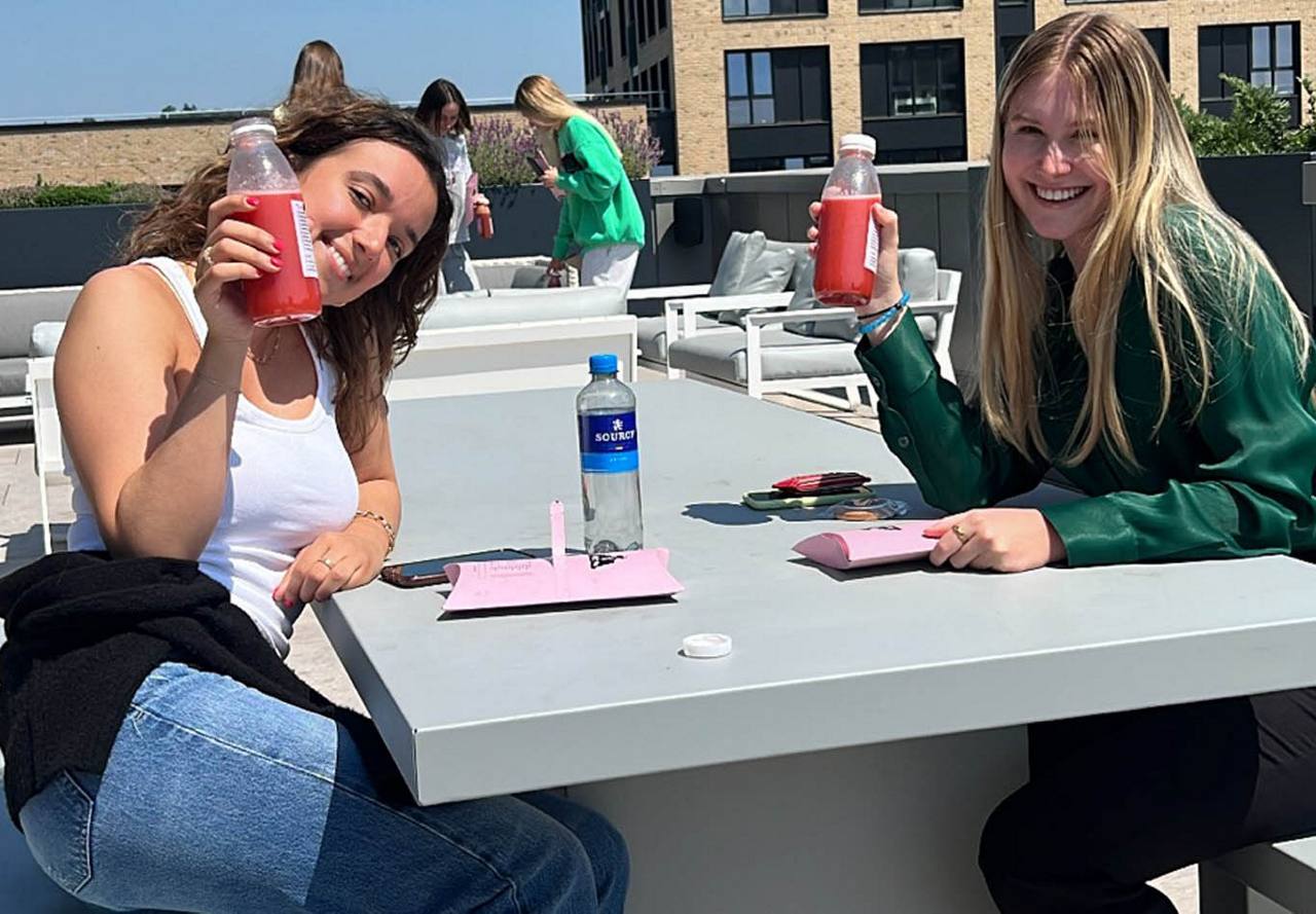 Two women smiling and toasting with bottles of juice while sitting at a table on a sunny rooftop terrace.