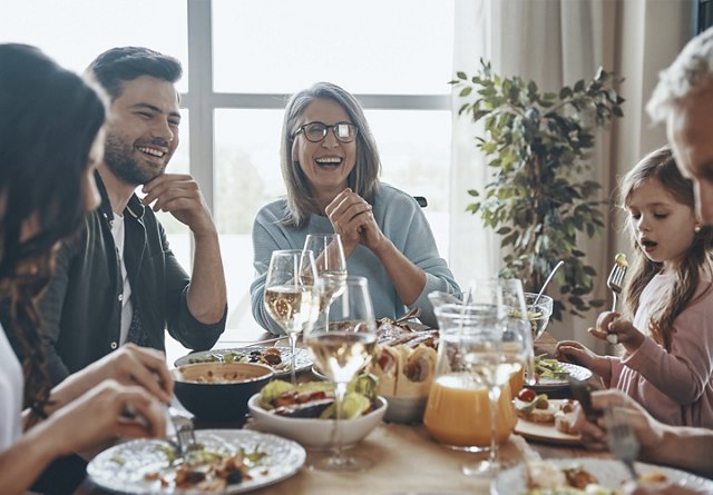 A group of people laughing and enjoying a meal together at a table with glasses of wine and various dishes.