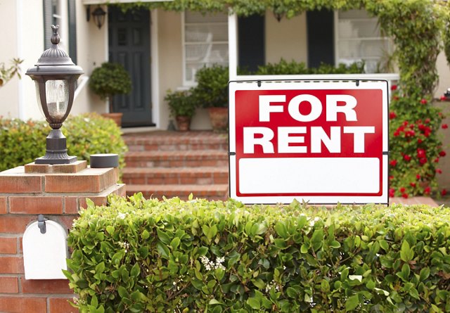 Red 'For Rent' sign posted in front of a residential home with a brick walkway, neatly trimmed hedge, and a classic lantern post.