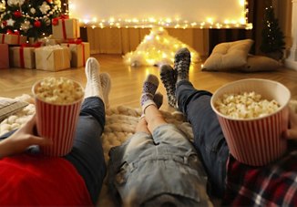 Two adults and a child relaxing indoors with their feet up, each holding a popcorn container, in a cozy room lit with Christmas lights and decorated with a tree and gifts.