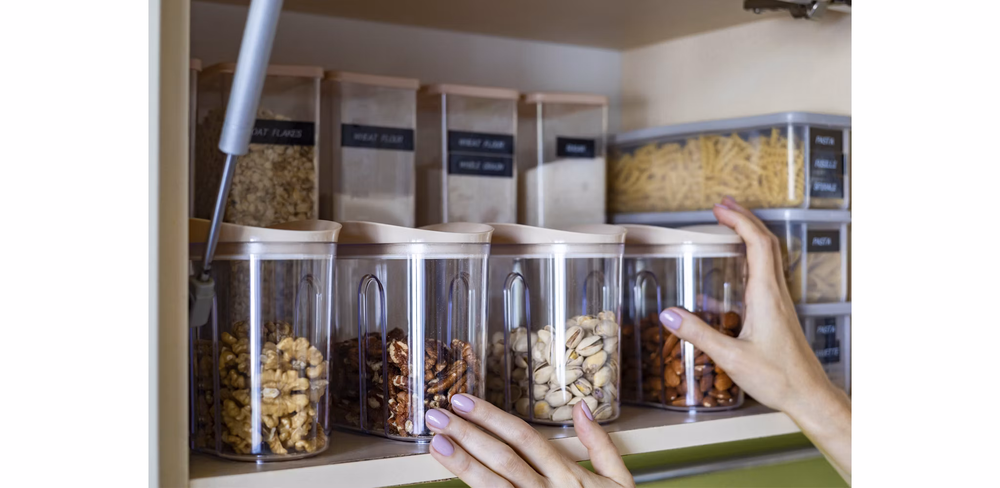 Hands organizing clear storage containers filled with various nuts and grains on a kitchen shelf.