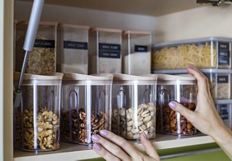 Hands organizing clear storage containers filled with various nuts and grains on a kitchen shelf.