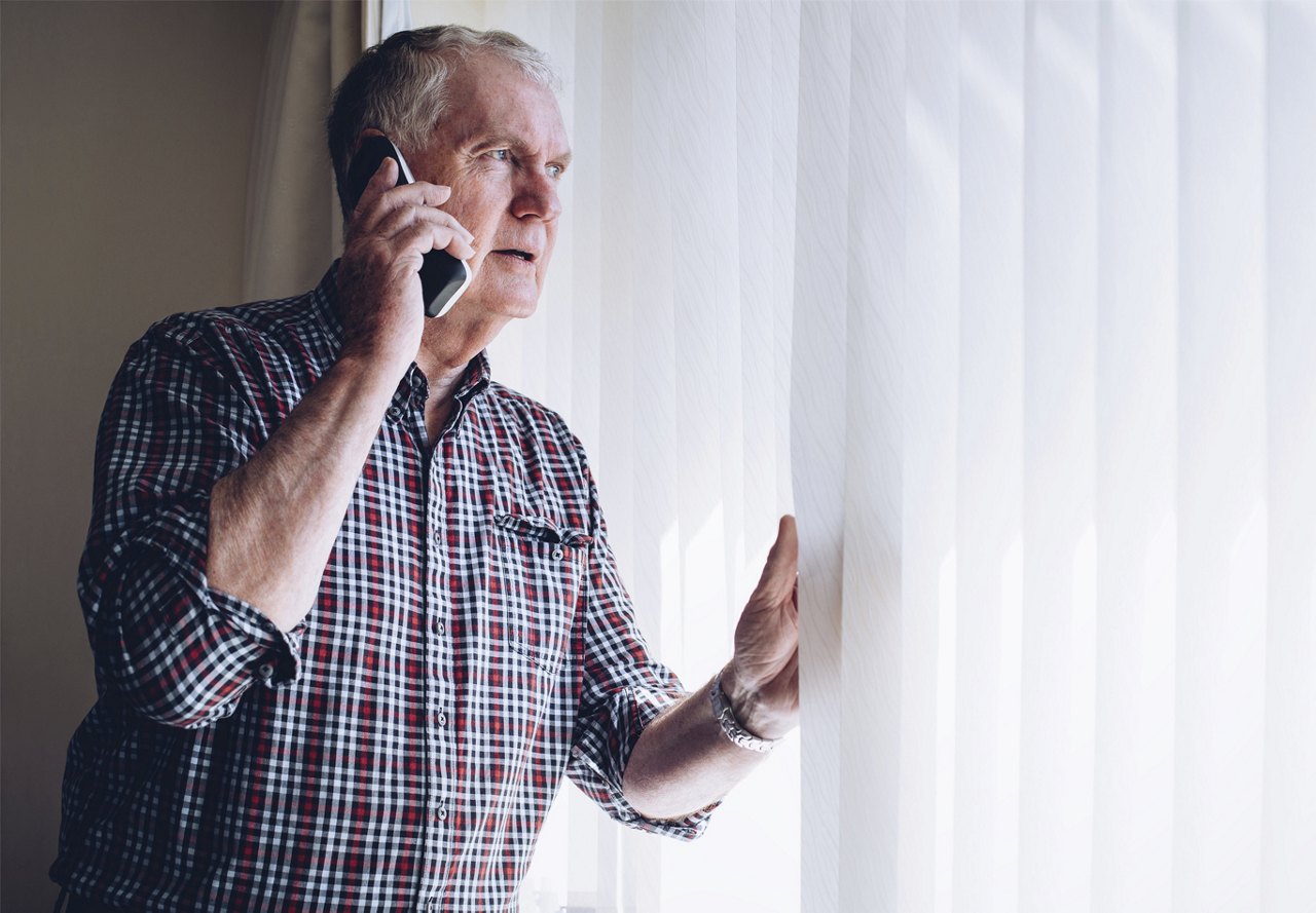 An elderly man in a plaid shirt talking on a mobile phone by a window with vertical blinds.