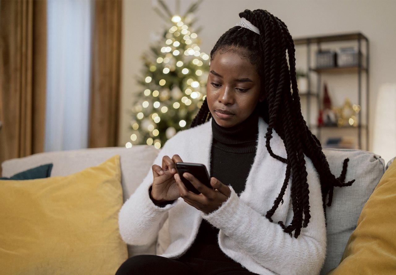 A woman with braided hair looking at her smartphone with a Christmas tree in the background.