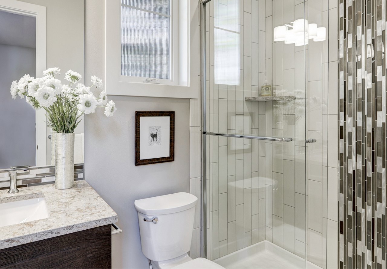 A contemporary bathroom with a glass shower, vase of white flowers, and framed artwork above the toilet.