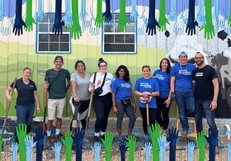 Group of volunteers wearing 'Better Together' t-shirts, smiling in front of a colorful mural with painted hands, holding gardening tools.