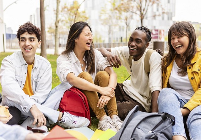 Four joyful students sitting on the grass at a park, laughing and enjoying each other's company, with backpacks suggesting they are on a break from classes.