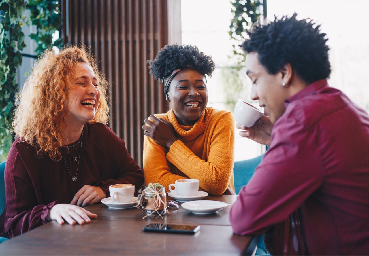 Three friends laughing and enjoying coffee together at a cafe table.