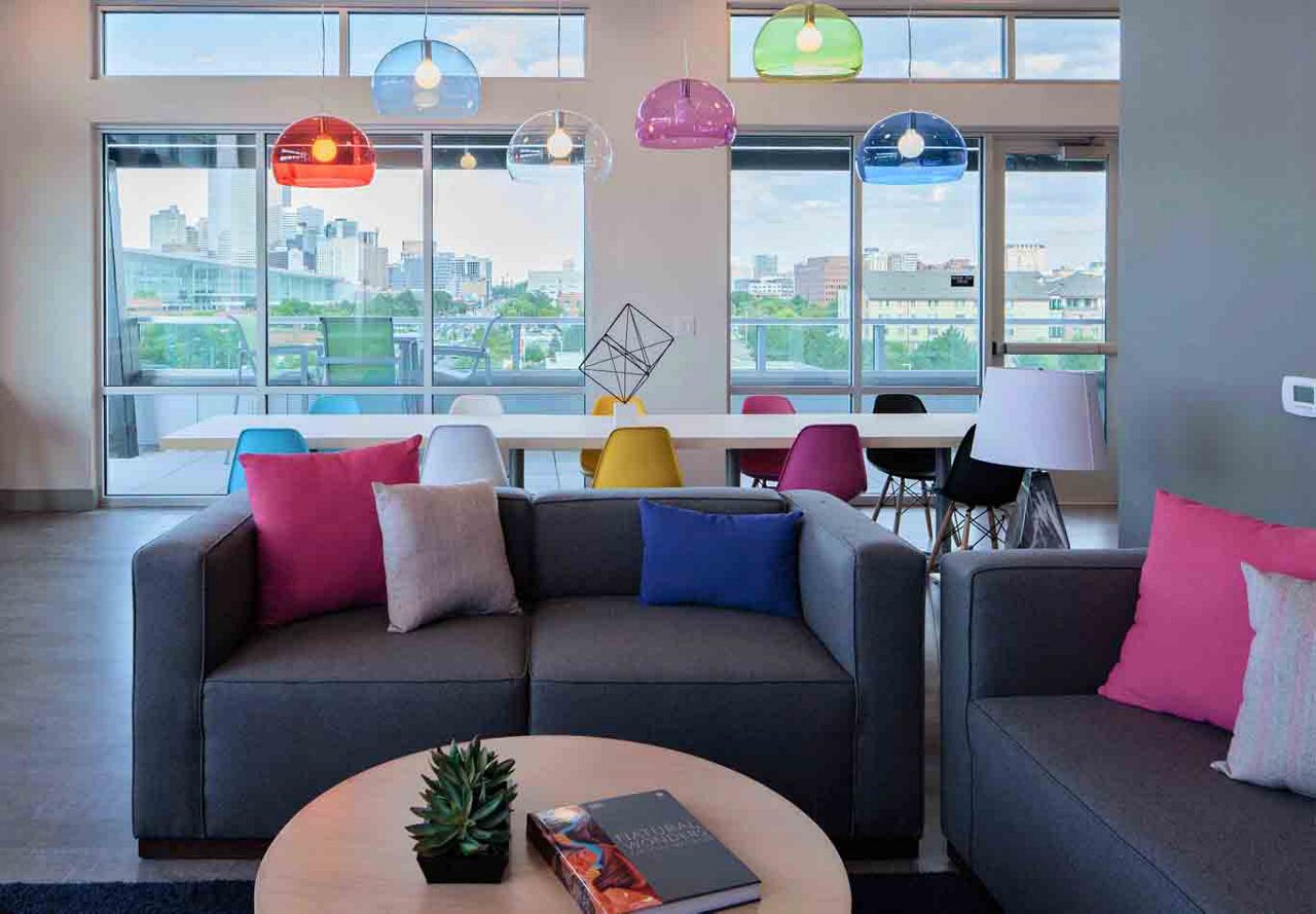 Modern common area with colorful furniture and hanging lamps, overlooking a cityscape through large windows.