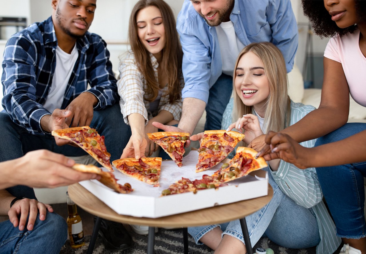 A diverse group of friends sharing slices of pizza from a box, sitting together with joyful expressions.