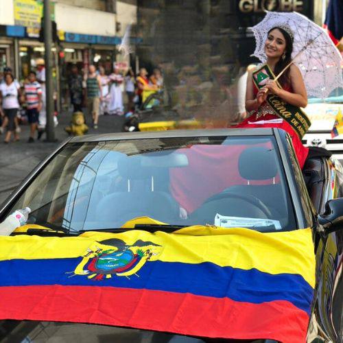 Beauty pageant participant waving from a car adorned with an Ecuadorian flag during a parade.