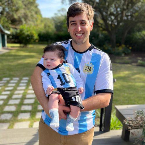 A man smiling and holding a baby, both wearing matching Argentina national football team jerseys.