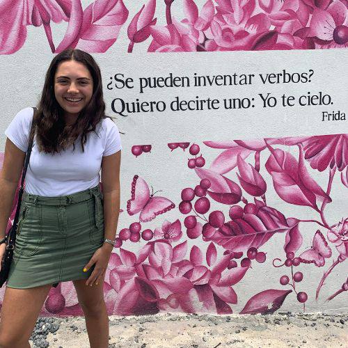 Patty Castillo, Investment Analyst, standing in front of a mural with pink floral patterns and a quote by Frida Kahlo in Spanish.
