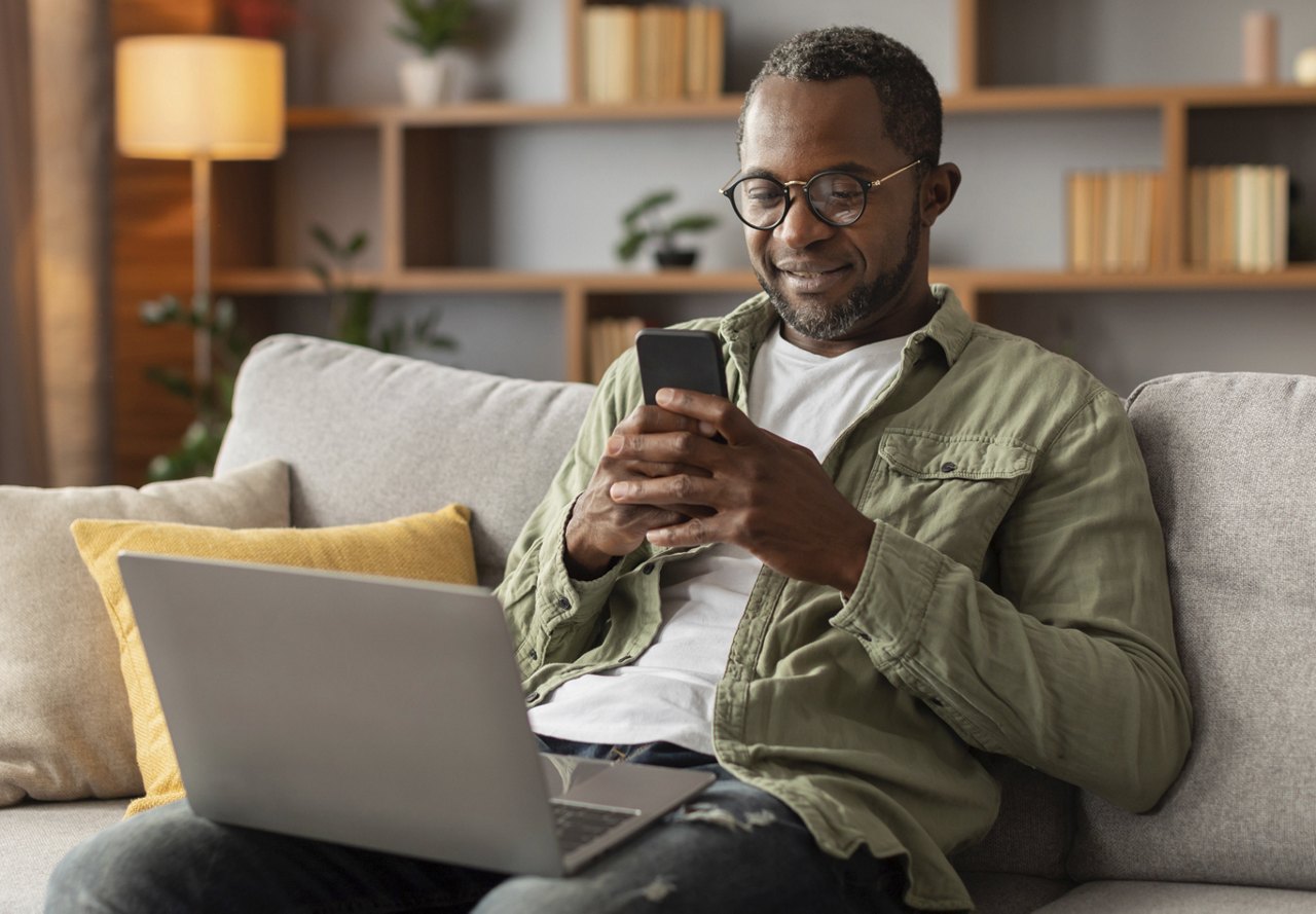 Man smiling while using a smartphone, with a laptop on his lap, comfortably seated on a couch at home.