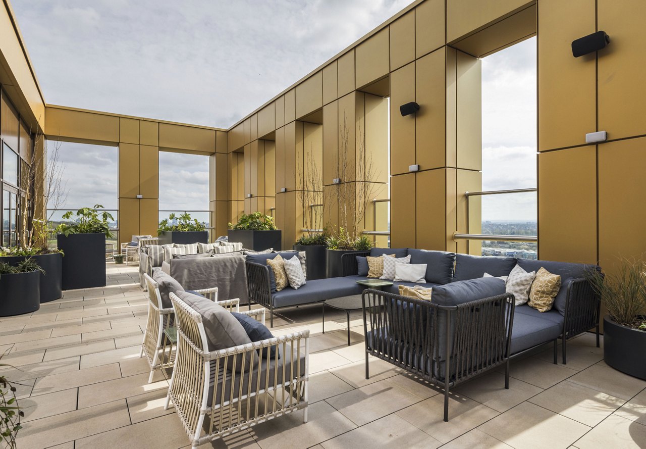 Rooftop lounge area with contemporary furniture, potted plants, and gold-paneled walls, offering a view of the horizon.